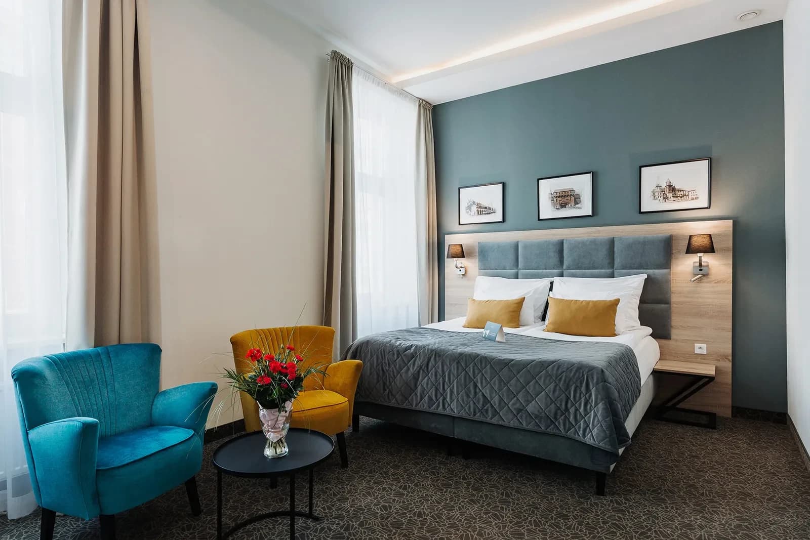 Contemporary bedroom corner with gray bed, teal and yellow armchairs, vase of red flowers.