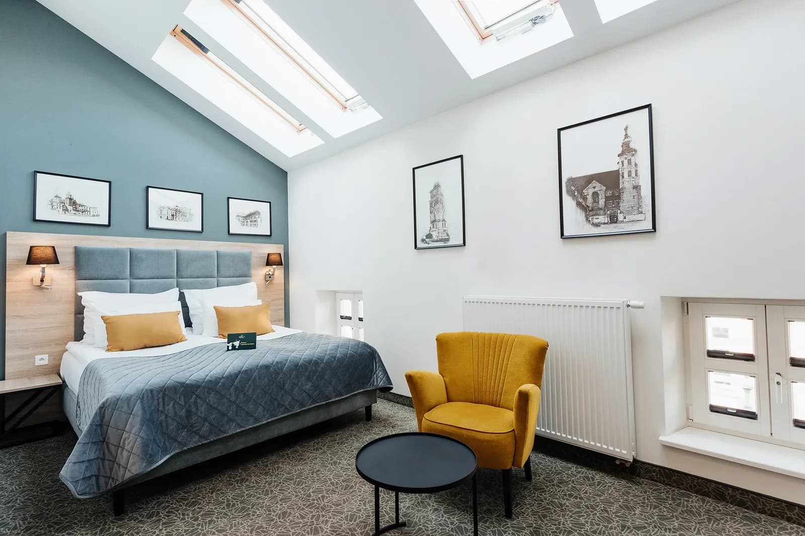 Attic bedroom with three skylights, gray quilted bed, mustard armchair and black round table.