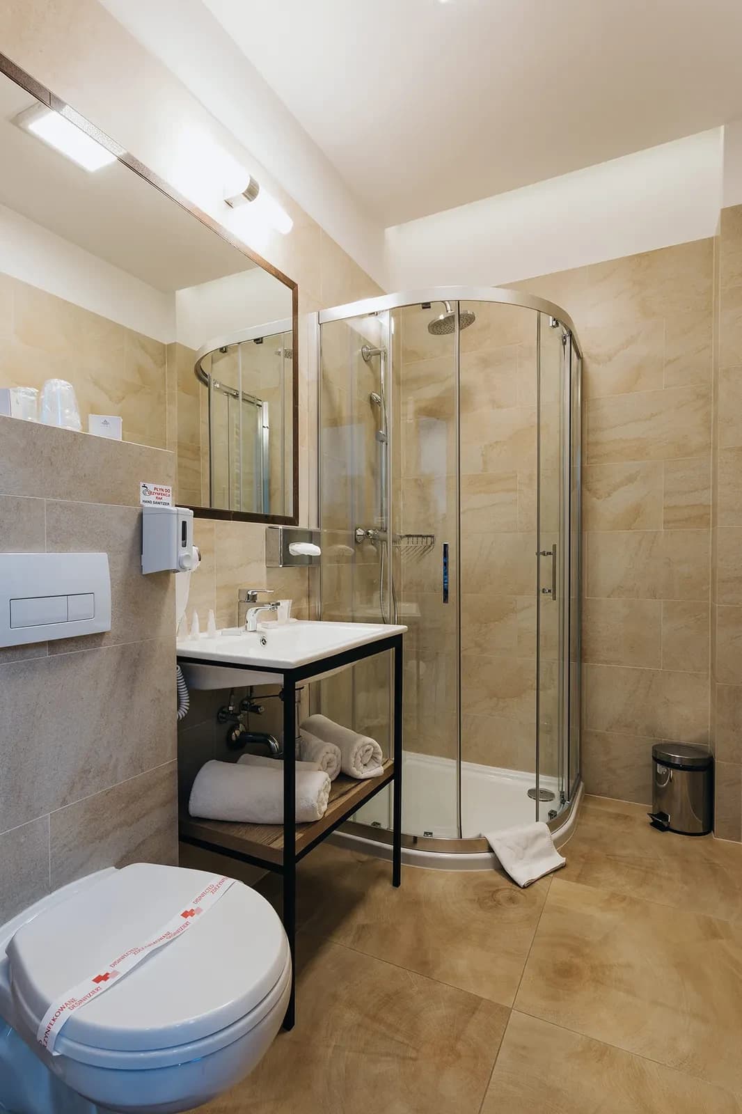 Small beige-tiled bathroom with glass shower enclosure, sink, rolled towels and sealed toilet.
