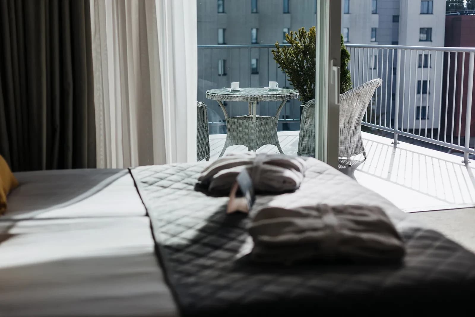 Bed in foreground opening to balcony with wicker table, two chairs, coffee cups, urban view.